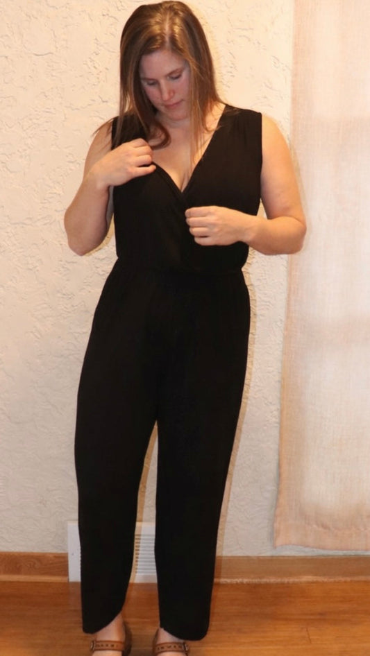 Woman wearing a black sleeveless jumpsuit standing against a plain wall.